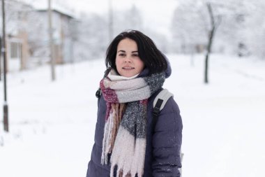 Genç, gülümseyen, güzel bir kadın kar yağarken açık havada fular takıyor.