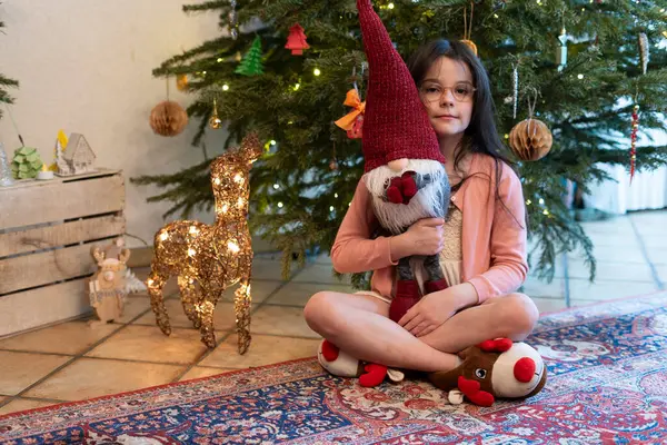 stock image Little cute girl in glasses hugging a Christmas gnome near a Christmas tree. Happy children's party concept