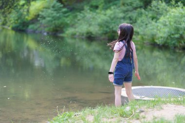 Little preschool girl in glasses and denim overalls near the pond. Outdoor childhood concept clipart