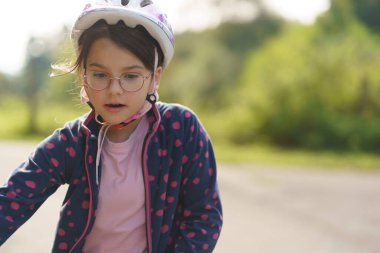 Little girl with glasses in helmet on the side of bike path. Concept of children's fun clipart