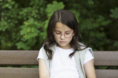 Gözlüklü, omuzlarında süveterle bankta oturan tatlı, üzgün bir kız. Hüzünlü çocukluk kavramı, sigorta, çocuk sağlığı.