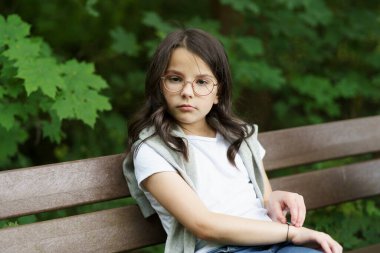 Gözlüklü, omuzlarında süveterle bankta oturan tatlı, üzgün bir kız. Hüzünlü çocukluk kavramı, sigorta, çocuk sağlığı.