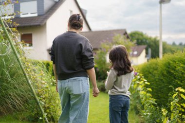 cute smiling brunette girl with pinned up hair in sweatshirt and jeans standing in front of her house on the lawn in the backyard with her little daughter dressed in the same way. Happy family life concept clipart