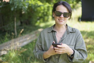Koyu renk saçlı, güneş gözlüklü ve yarım düğmesiz gömlekli yaz çiftliği arka planında poz veren elinde cep telefonu, dişinde çim sapı olan bir kız. Tarım, güzellik, mobil teknolojiler kavramı