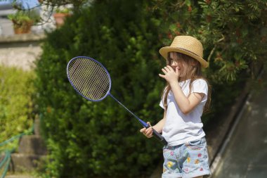 Hasır şapkalı ve bir dişi eksik şirin küçük kız yazın bahçede badminton oynuyor. Neşeli, sağlıklı, aktif bir çocuk kavramı