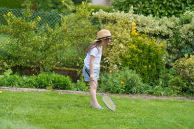 Hasır şapkalı ve bir dişi eksik şirin küçük kız yazın bahçede badminton oynuyor. Neşeli, sağlıklı, aktif bir çocuk kavramı