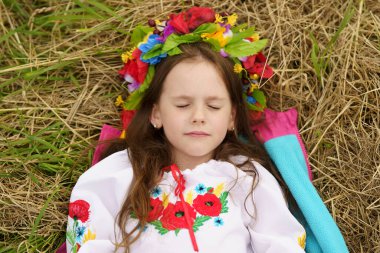 A little beautiful girl in a wreath and embroidered shirt lying on a freshly mown field. The concept of a happy childhood and traditions in the bosom of nature clipart