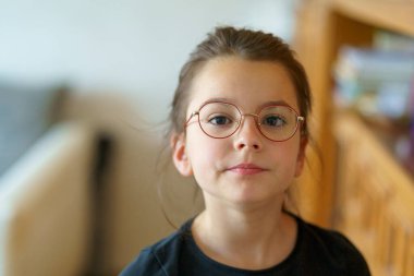 little cute girl with glasses standing near big window. Smart conscious child concept clipart