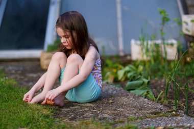 Little sad lightly dressed wet girl sits on concrete path in backyard in summer. Concept of difficult childhood clipart