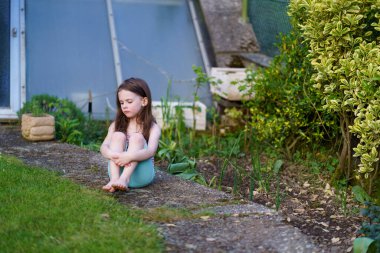 Little sad lightly dressed wet girl sits on concrete path in backyard in summer. Concept of difficult childhood clipart