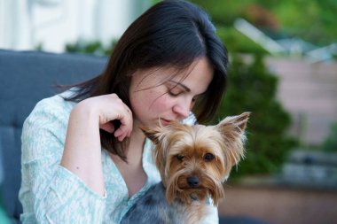 Cute dark-haired girl dressed lightly smiling sits with her small dog at a table in a cafe in the summer. Yorkshire terrier in the arms of its owner clipart