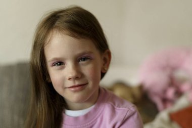 Göz makyajlı, pembe kazaklı, gülümseyen güzel bir kız kanepeye oturur ve arka planı bulanıktır. Sağlıklı neşeli aktif çocuk konsepti