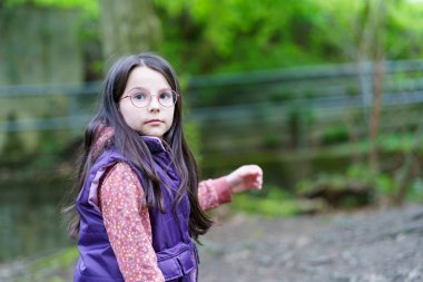 Küçük, güzel, gülümseyen, uzun saçlı, gözlüklü ve yelekli bir kız ormanda tek başına duruyor.