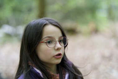 Küçük, güzel, gülümseyen, uzun saçlı, gözlüklü ve yelekli bir kız ormanda tek başına duruyor.