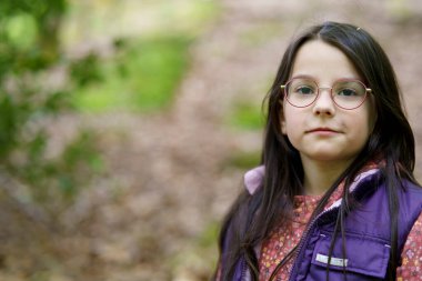 Küçük, güzel, gülümseyen, uzun saçlı, gözlüklü ve yelekli bir kız ormanda tek başına duruyor.