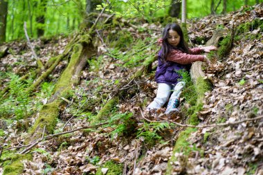 Küçük, güzel, gülümseyen, uzun saçlı, gözlüklü, siyah saçlı bir kız ormanda yelek giyiyor.