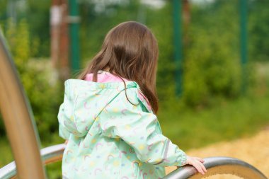 Lonely sad girl on the playground in a raincoat. Concept of difficult childhood and psychological problems clipart