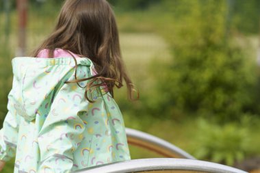 Lonely sad girl on the playground in a raincoat. Concept of difficult childhood and psychological problems clipart