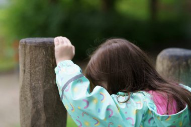 Lonely sad girl on the playground in a raincoat. Concept of difficult childhood and psychological problems clipart