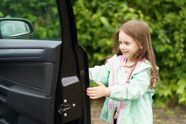 Little cheerful girl in raincoat opens door of black car. Concept of children's travel, fun clipart