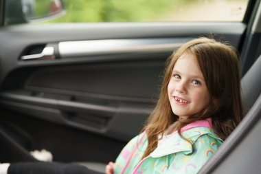 Yağmurluk giymiş, gülümseyen neşeli kızıl saçlı kız arabada oturuyor. Çocuk seyahati, eğlence, güvenlik kavramı