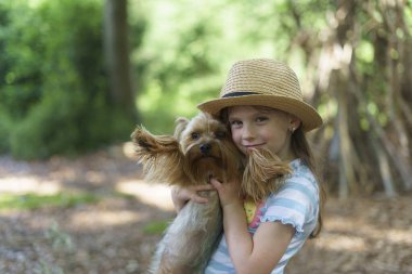 Hasır şapkalı küçük bir kız yaz ormanında küçük bir köpeğe sarılıyor. Çocuk ve hayvan dostluğu kavramı