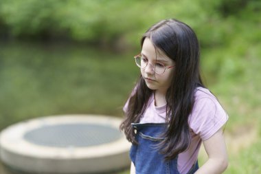 Gözlüklü ve kot pantolonlu küçük bir kız yazın bir yaz parkında gölet kenarında oynuyor. Açık havada mutlu ve aktif bir çocuk kavramı