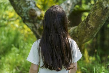 Ormanda bir ağacın altında duran beyaz tişörtlü genç bir kızın arka görüntüsü.