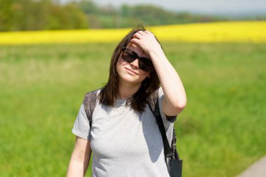 Güneş gözlüğü takan genç bir kadın arka planda ağaçlarla çayırda yürüyor. Etkin seyahat konsepti