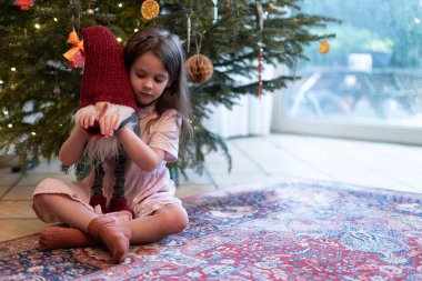 A little sad beautiful girl sits dressed up under a Christmas tree hugging a Christmas gnome. a deer from a garland stands nearby clipart