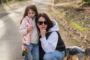 Mother and daughter dressed in vests hugging on a path in a spring forest. Concept of warm family relations clipart