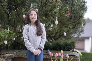 Little cute girl in glasses with long hair smiling standing near Easter tree. Concept of children's joy and religion clipart