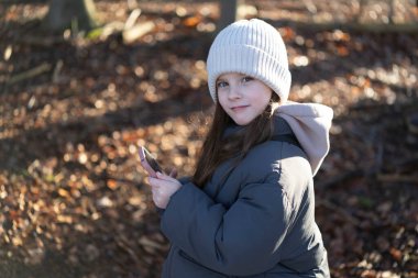Küçük, şirin, uzun saçlı, şapkalı bir Alman kızı elinde akıllı bir telefonla bir sonbahar ormanının ortasında duruyor. Doğaya çocuk gezisi kavramı