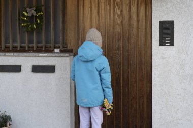 A little teenage girl in a jacket and hat stands with her back to the camera near the door of her house. Concept of childhood loneliness clipart