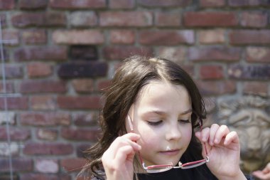Küçük güzel bir kız gözlüğünü tuğla bir duvara dayamış.