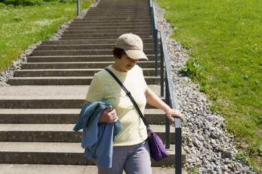 Yaşlı bir kadın elinde tırabzan tutarak yaz parkında merdivenlerden iniyor. Yaşlılık konseptinde aktif yaşam