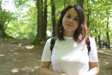 Cute wide-eyed girl in a T-shirt stands smiling in the middle of a summer park. Good mood concept clipart