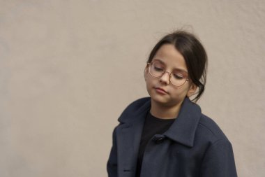 A young girl with light skin, wearing round glasses and a dark blue coat, stands against a beige wall with a pensive expression. The image represents loneliness, deep thoughts, or childhood melancholy. clipart