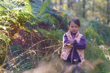 Kapüşonlu ve yelekli güzel bir kız bir sonbahar ormanının arka planında duruyor. Doğada mutlu bir çocukluk kavramı.