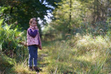 Kapüşonlu ve yelekli güzel bir kız bir sonbahar ormanının arka planında duruyor. Doğada mutlu bir çocukluk kavramı.