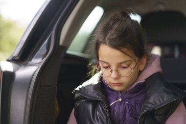 Gözlüklü ve yelekli küçük üzgün bir kız ormanda yürüdükten sonra arabanın bagajında yatıyor..