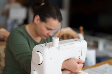 Tişört giyen, arka planı bulanık bir odada dikiş makinesine dikiş diken tatlı, güzel bir kız. Ev yaratıcılığı kavramı.