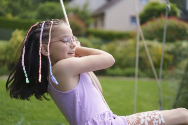 Küçük sevimli bir gülümseme ve gözlüklü pozitif bir kız ve renkli örgülü bir elbise yeşil ve çiçeklerle çevrili bir yaz bahçesinde sallanıyor. Sağlıklı bir çocuk ve duygu kavramı.