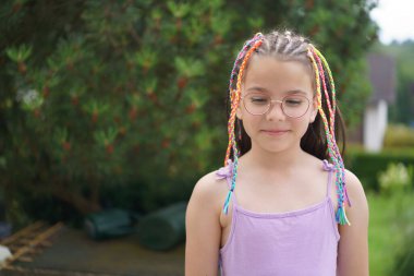 Küçük sevimli bir gülümseme, gözlüklü pozitif bir kız ve yaz bahçesinde yeşillik ve çiçeklerle çevrili renkli örgülü bir elbise. Sağlıklı bir çocuk ve duygu kavramı.