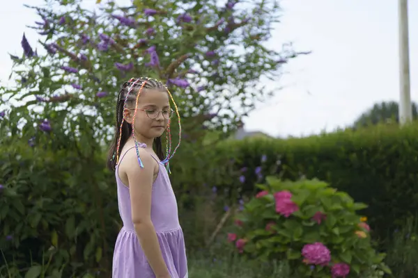 Küçük sevimli bir gülümseme, gözlüklü pozitif bir kız ve yaz bahçesinde yeşillik ve çiçeklerle çevrili renkli örgülü bir elbise. Sağlıklı bir çocuk ve duygu kavramı.