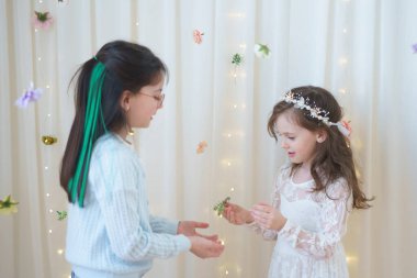 Perdenin önünde duran iki kız. Beyaz elbiseli ve taç takan bir kız.