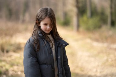 Dışarıda ormanda bir patikada ceketini giymiş kameraya bakan bir kız. Çocukların boş vakitleri ve duyguları kavramı