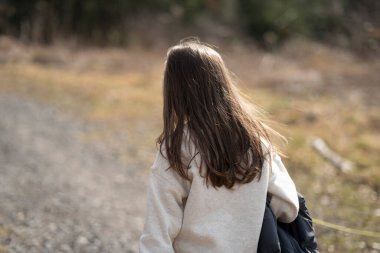 Sırtı kameraya dönük ve elinde ceketi olan bir kız ormanda bir patikada kesik ağaç gövdesi yığınına karşı. Çocukların yalnızlığı ve duyguları kavramı