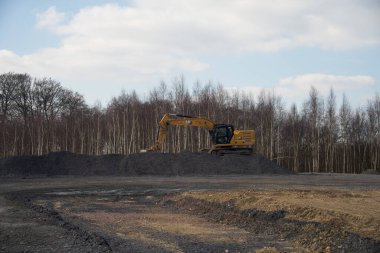 Iserlohn, Kuzey Ren-Vestfalya, Almanya. 16 Mart 2025. Ağaçların önündeki çakıl yığınının üzerinde sarı bir kazıcı. İnşaat konsepti.