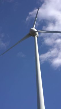 Tepenin üzerinde beyaz bulutlu mavi bir gökyüzüne karşı yalnız bir rüzgar türbininin 4k dikey videosu. Yenilenebilir enerji ve sürdürülebilir kalkınma kavramı